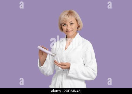 Belle femme mature en peignoir avec bouteille de produit cosmétique sur fond violet Banque D'Images