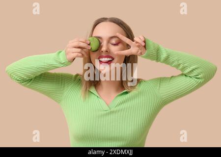 Belle jeune femme avec macaron vert doux montrant un geste de victoire sur fond beige Banque D'Images