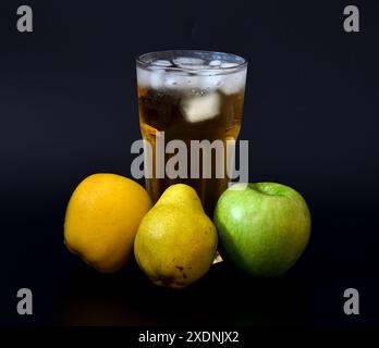 Jus de pomme et de poire avec de la glace dans un grand verre à facettes sur un fond noir, avec des fruits mûrs à proximité. Gros plan. Banque D'Images