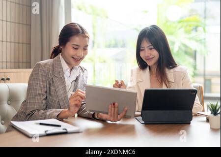 Deux jeunes femmes d’affaires asiatiques positives discutent de leur travail sur leurs tablettes numériques, ont une réunion informelle et travaillent ensemble dans un café Banque D'Images