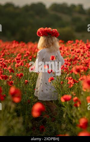 Une jeune fille portant une couronne de fleurs rouges marche à travers un champ de coquelicots rouges. La scène est sereine et paisible, avec la fille se démarquant contre le Banque D'Images