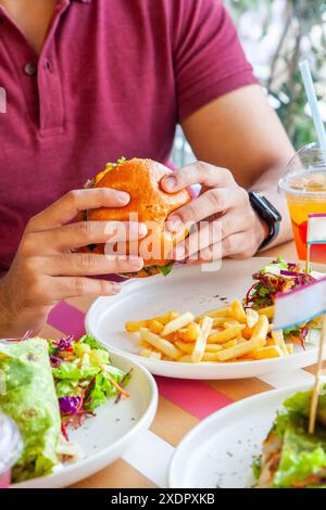 Garçon homme tenant un burger de poulet frit glacé au miel dans un pain brioché servi avec des frites et une salade d'accompagnement sur une assiette dans un environnement de café. Banque D'Images