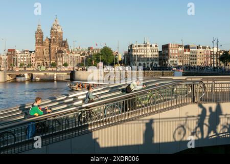 Les cyclistes poussent leurs vélos vers le haut du Travelator depuis le parking souterrain de la gare centrale, avec le canal et la basilique Saint-Nicolas en arrière-plan, à Amsterdam, aux pays-Bas. Banque D'Images