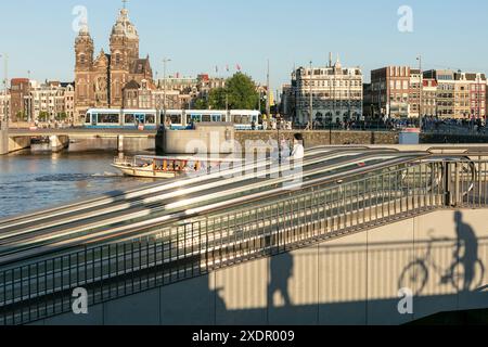 Une cycliste pousse son vélo sur le Travelator jusqu’au parking souterrain de la gare centrale, avec le canal et la basilique Saint-Nicolas en arrière-plan, à Amsterdam, aux pays-Bas. Banque D'Images