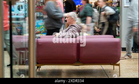 Photographie longue vitesse d'obturation d'un homme plus âgé assis sur un siège dans un mal de shopping, avec des acheteurs se déplaçant autour et derrière lui Banque D'Images