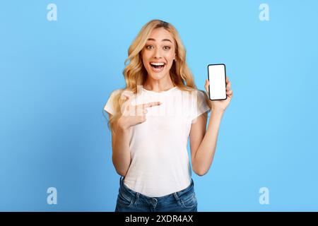 Excité jeune femme tenant smartphone avec écran vierge contre le bleu Banque D'Images