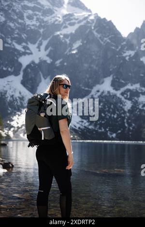 Une fille voyageuse avec un sac à dos profite de la vue imprenable sur une chaîne de montagnes et un lac, avec des sommets enneigés en arrière-plan Banque D'Images