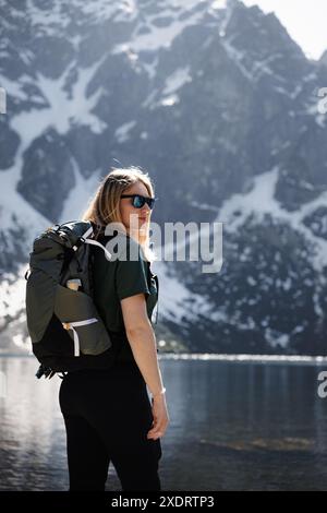 Une fille voyageuse avec un sac à dos profite de la vue imprenable sur une chaîne de montagnes et un lac, avec des sommets enneigés en arrière-plan. Banque D'Images