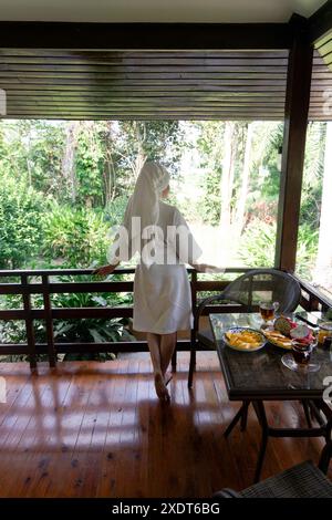 Femme dans une robe blanche se tient sur un balcon donnant sur une végétation tropicale luxuriante, profitant de la vue. Banque D'Images