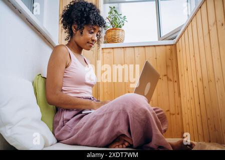 La femme est assise confortablement dans un coin confortable avec des murs en bois, travaillant sur son ordinateur portable, créant un environnement détendu et productif. Travail en ligne à distance à domicile. Banque D'Images