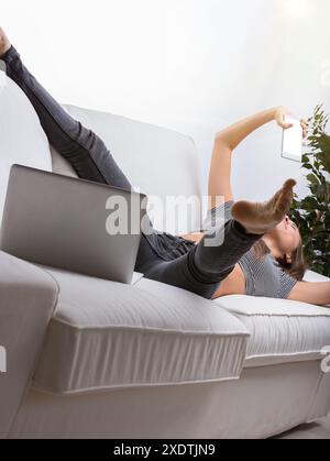 Jeune femme se détendant sur son canapé à la maison, en utilisant son smartphone et en prenant un selfie dans un salon élégant. Profitez d'une journée de repos à l'intérieur Banque D'Images