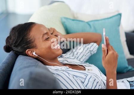 Femme, smartphone et souriant sur le canapé avec écouteurs, radio et application mobile pour l'audio sur le week-end pause dans la maison. Écran, visionnage et contenu vidéo activé Banque D'Images