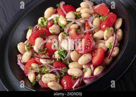 Piyaz, salade de haricots blancs turcs avec tomates, oignon rouge et persil aromatisée au sumac et vinaigrette à l’huile d’olive dans un bol noir sur une table en bois noir, Banque D'Images