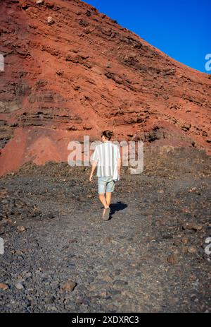 Un personnage solitaire marche à travers un paysage rocheux et volcanique, baigné de lumière chaude du soleil. Les falaises rouges en arrière-plan créent un contraste frappant avec le Banque D'Images