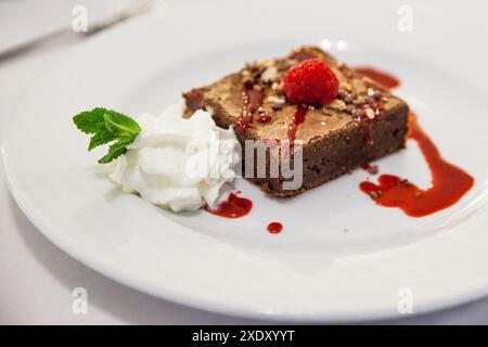 un morceau de gâteau au chocolat et aux noisettes servi sur une assiette blanche dans un restaurant décoré de crème fouettée, menthe et framboises Banque D'Images