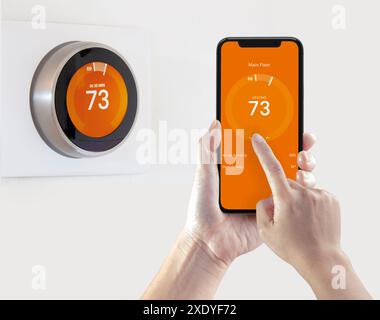 Une personne tenant un iPhone 11 Pro Max à côté d'un thermostat intelligent Nest pendant que la chaleur est allumée. Banque D'Images