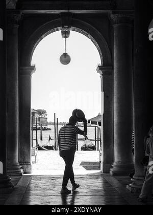 Venise, Italie - 30 juin 20220 touriste marche sous une arche à venise, italie, tenant son chapeau pendant que les gondoles attendent en arrière-plan Banque D'Images