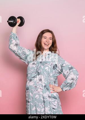 Happy young pregnant woman wearing blue dress faire de l'exercice d'haltères de levage. Studio shot sur fond rose. Mode de vie sain et de remise en forme au cours de pr Banque D'Images