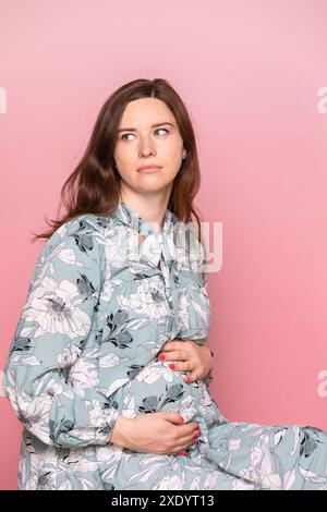Pensive jeune femme enceinte portant une robe florale tenant ses mains sur son ventre. Studio tourné sur fond rose avec espace de copie Banque D'Images