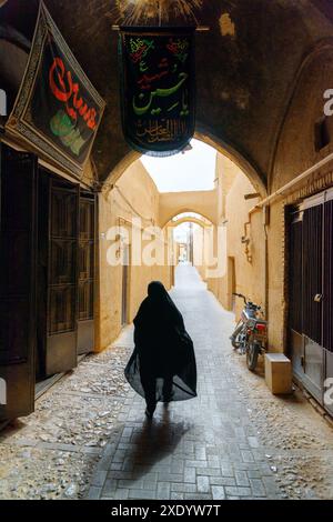 Yazd, Iran - 26 octobre 2018 : femme iranienne portant un tchador noir marchant le long d'une rue étroite de la ville historique de Yazd. Banque D'Images