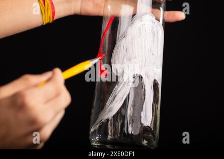 Un artiste tenant et peignant une bouteille en verre avec de la gouache rouge et blanche noire. Isolé sur fond noir. Banque D'Images