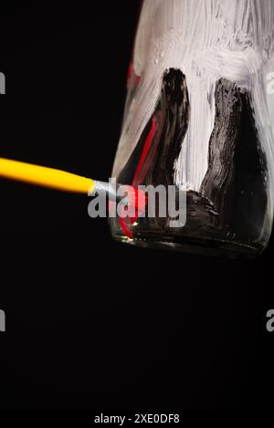 Une bouteille en verre étant peinte avec un pinceau dans les couleurs blanc, rouge et noir. Isolé sur fond noir. Banque D'Images