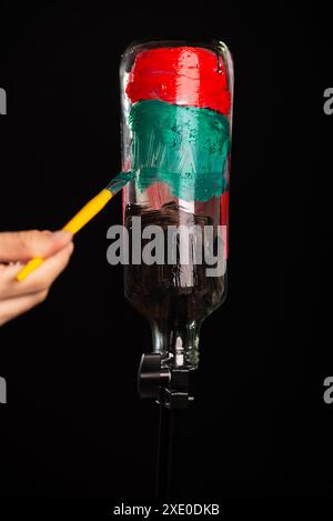 Une main de peintre peint une bouteille en verre avec des peintures. Isolé sur fond noir. Banque D'Images