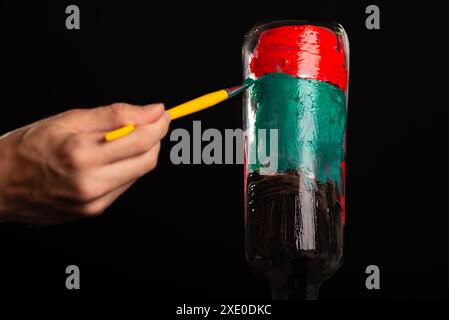 Une main de peintre peint une bouteille en verre avec des peintures. Isolé sur fond noir. Banque D'Images