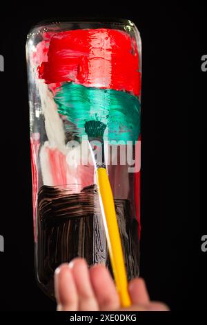 Une main de peintre peint une bouteille en verre avec des peintures. Isolé sur fond noir. Banque D'Images