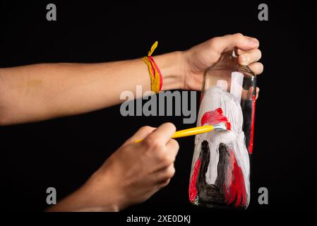 Un artiste tenant et peignant une bouteille en verre avec de la gouache rouge et blanche noire. Isolé sur fond noir. Banque D'Images