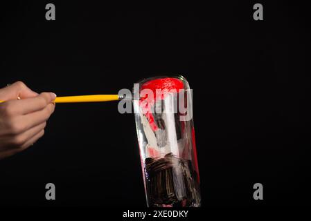 Une bouteille en verre étant peinte avec un pinceau dans les couleurs blanc, rouge et noir. Isolé sur fond noir. Banque D'Images