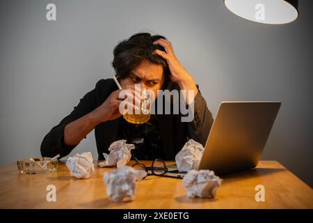 Un homme boit un verre de bière assis à une table avec un ordinateur portable et une pile de papier. La scène suggère une atmosphère décontractée et détendue Banque D'Images