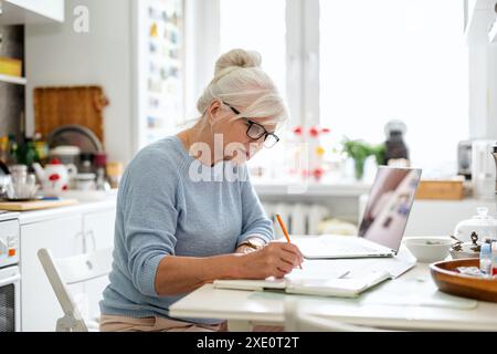 Femme mature utilisant un ordinateur portable tout en étant assis à la table dans la cuisine Banque D'Images