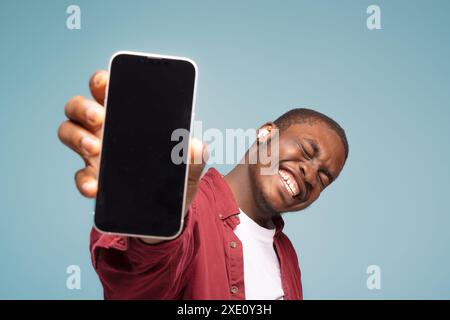 Publicité mobile. Homme noir ravi pointant vers le téléphone portable avec écran blanc vide sur fond bleu de studio, maquette pour l'application ou le site Web. Cellphon Banque D'Images
