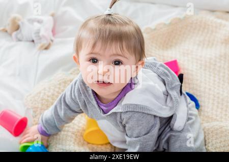 Bébé fille dans un pull gris rampant heureusement sur un lit entouré de tasses colorées, explorant avec joie et curiosité. L'image capture l'innoce de l'enfance Banque D'Images