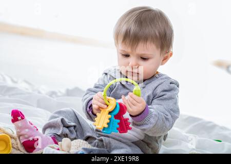 Bébé fille est assis sur une couverture blanche et joue avec un ensemble de clés en plastique colorées. Le bébé se concentre sur le jouet dans ses mains Banque D'Images