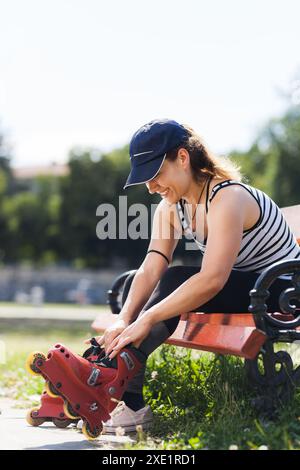 Jeune femme joyeuse profite d'une journée ensoleillée dans le parc alors qu'elle se prépare au roller. Portant une casquette et une tenue de sport, elle ajuste ses patins rouges. Banque D'Images