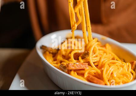 Gros plan d'une personne mangeant des spaghettis avec une fourchette d'un plat blanc. Les spaghettis sont recouverts d'une sauce orange crémeuse et garnis de petits morceaux Banque D'Images