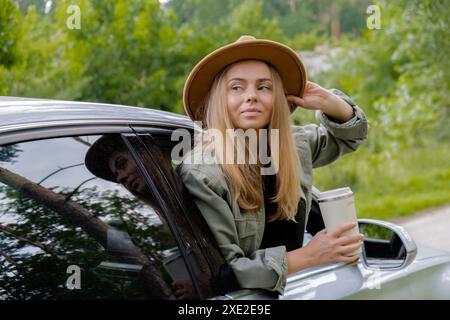 Femme blonde sortant la tête de la voiture de pare-brise et buvez du café ou du thé dans une tasse réutilisable. Les jeunes touristes explorent les voyages locaux m Banque D'Images