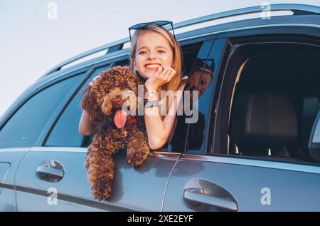 Portrait chien Maltipoo brun pelucheux avec une petite fille souriante regardant par une fenêtre de voiture ouverte alors qu'ils sont assis sur le siège arrière pendant le voyage en voiture. Fu Banque D'Images