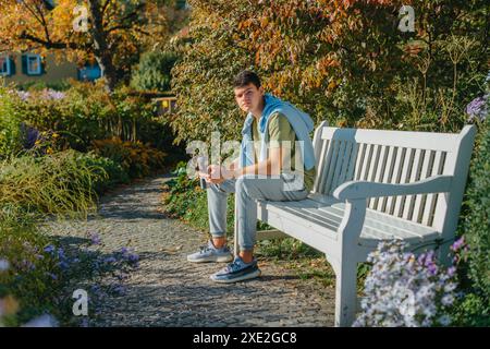 Un adolescent assis sur Un banc dans le parc d'automne boit du café dans Une tasse Thermo et regarde dans Un téléphone. Portrait de beau cheer Banque D'Images