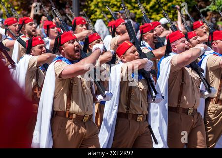 Oviedo, Espagne - 25 mai 2024 : des rangées de soldats chantants en uniformes militaires, casquettes rouges et capes marchent en formation avec des fusils tenus haut pendant un millier Banque D'Images