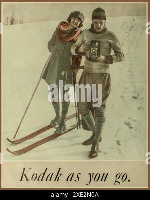 Publicité KODAK vintage des années 1920, mettant en vedette un couple sur la neige en hiver, intitulée « Kodak as You Go ». Homme tenant un appareil photo à film pliable Kodak à soufflets avec un câble de déverrouillage prenant une photo sur film Kodak. Le début de la photographie grand public populaire telle que nous la connaissons, à travers la vision de George Eastman. Banque D'Images