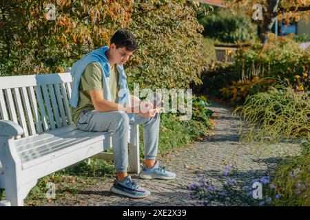 Un adolescent assis sur Un banc dans le parc d'automne boit du café dans Une tasse Thermo et regarde dans Un téléphone. Portrait de beau cheer Banque D'Images