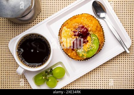 Vue de dessus de délicieux danoises de fruits mélangés maison, une tasse de café noir, des raisins et une cuillère en métal dans un plateau en plastique blanc. Banque D'Images