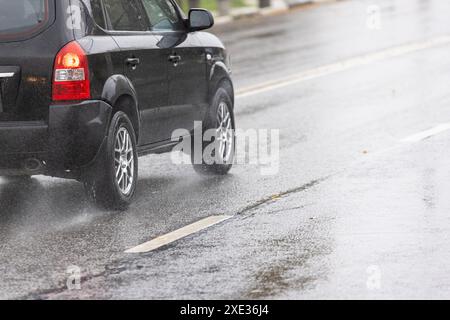 Les éclaboussures d'eau de pluie coulent des roues de SUV noir qui se déplacent rapidement sur la route asphaltée Banque D'Images