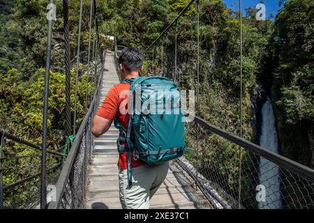 Jeune aventurier latin avec un sac à dos traversant un pittoresque pont suspendu en bois près de la majestueuse cascade de Pailon del Diablo à Baños de Agua San Banque D'Images