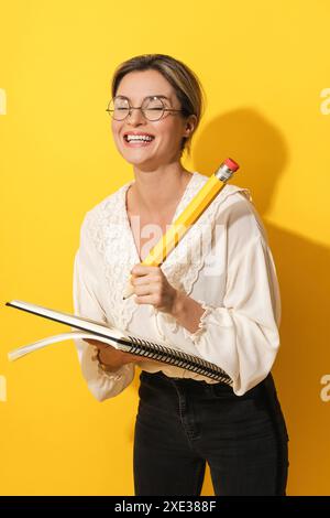 Femme joyeuse portant des lunettes tenant un grand crayon et un cahier sur fond jaune Banque D'Images