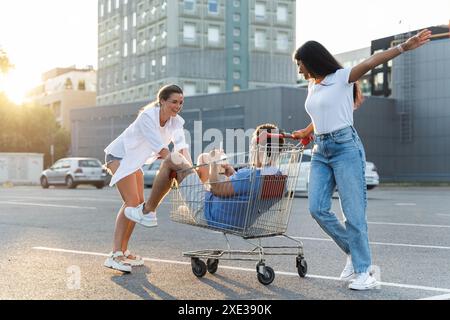 Trois amis diversifiés ayant du plaisir et le chariot d'équitation sur le parking près d'un supermarché Banque D'Images