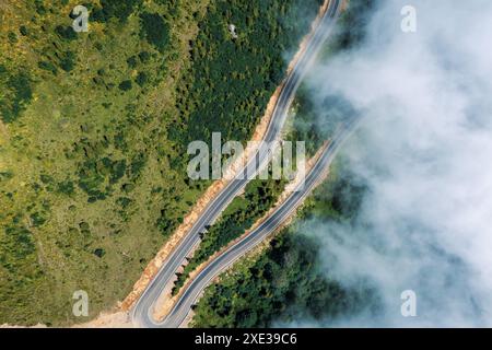 Vue aérienne de dessus de la courbe de montagne route sinueuse à travers un paysage verdoyant luxuriant avec la brume matinale et les nuages. La route serpentine traverse une forêt animée, offrant un itinéraire de voyage pittoresque. Banque D'Images